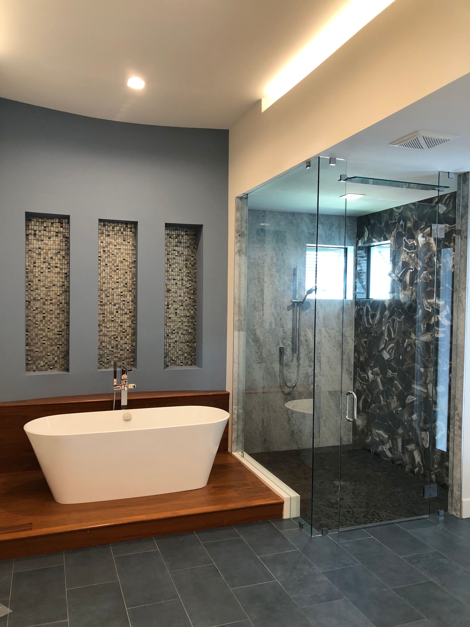 A modern bathroom featuring a white soaking tub on a wood platform against a blue wall with three tiled niches next to a shower.