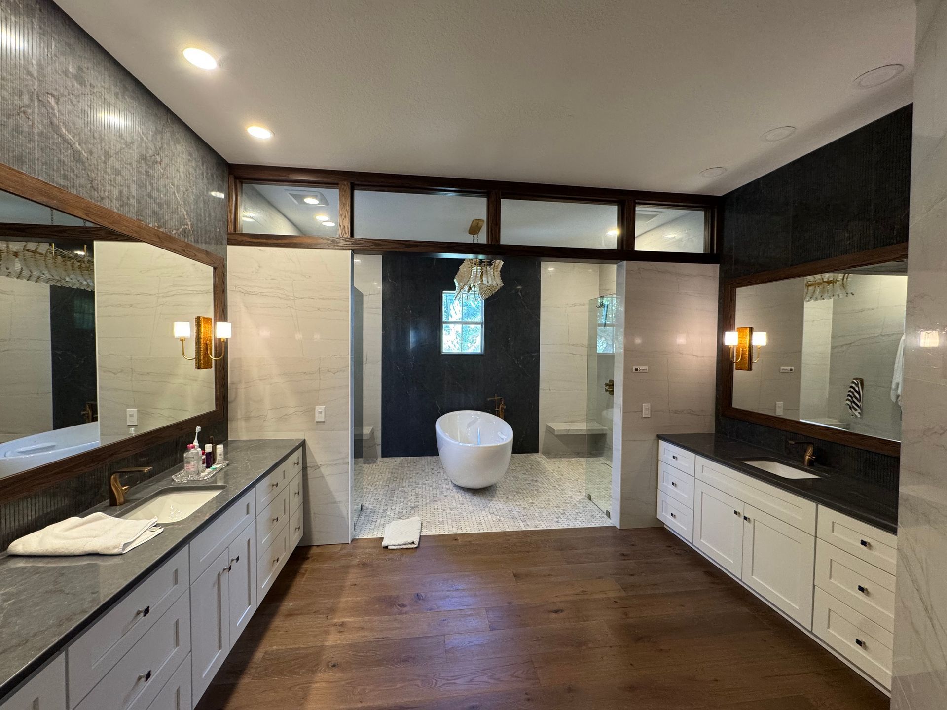A spacious modern bathroom with a white soaking tub, wood flooring, dual vanities with white cabinets, and large mirrors.