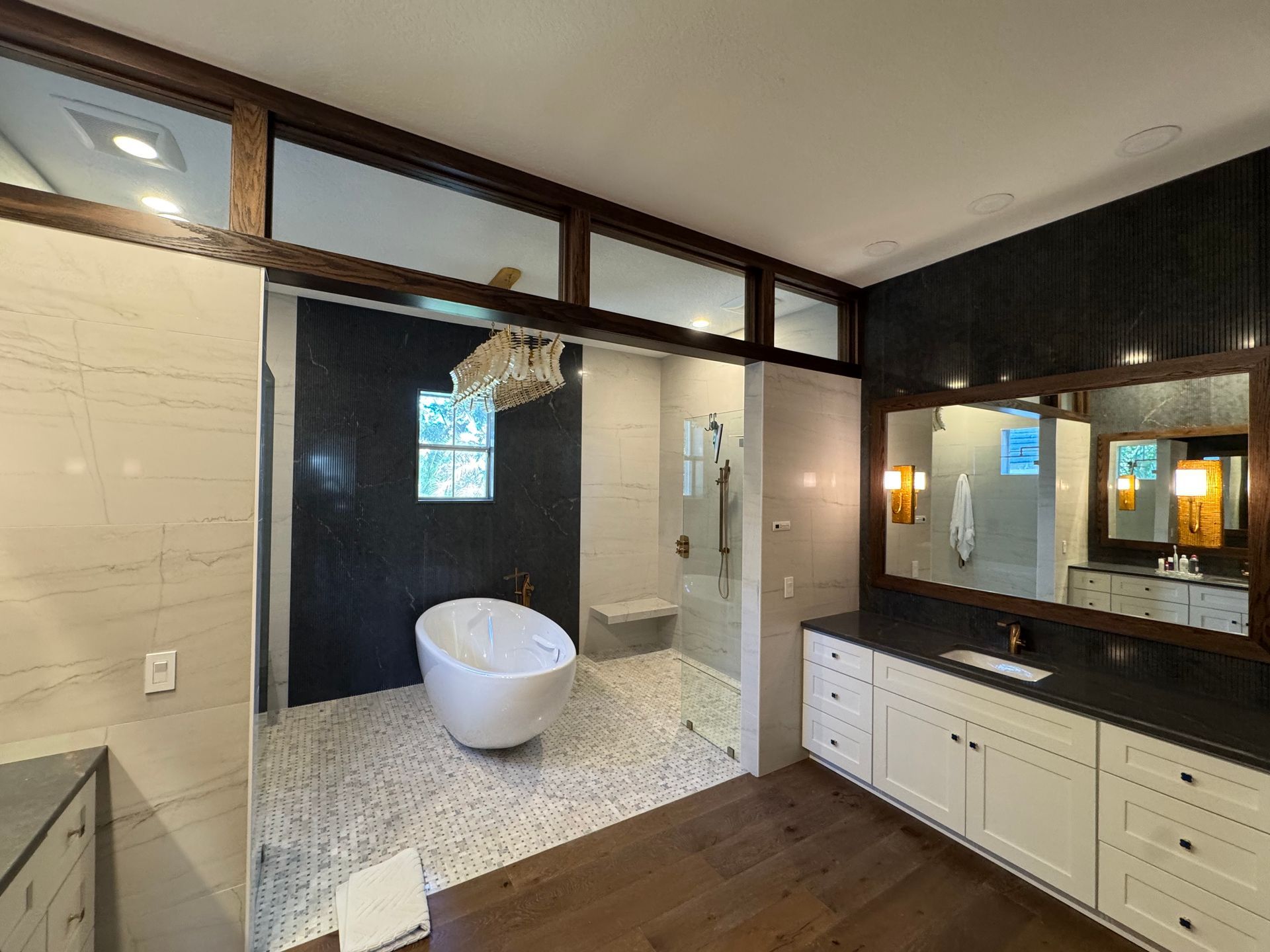 A modern bathroom featuring a white freestanding tub, glass-enclosed shower, dual-sink vanity, and hardwood flooring.