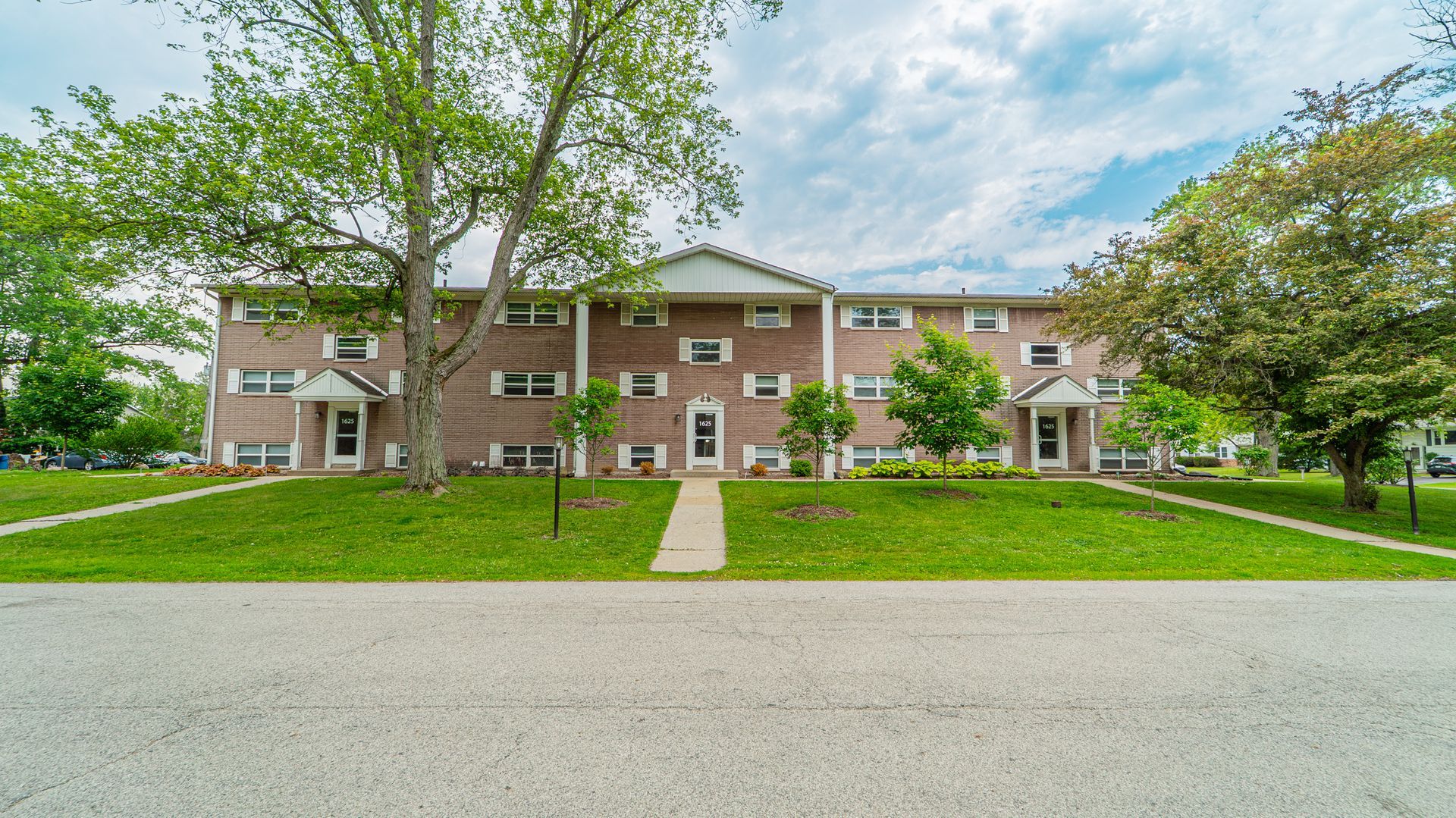 Row of two-story brick townhouses with green lawns, trees, and a concrete path leading to the doors.