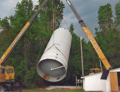 Two cranes lift a large, cylindrical metal tank in a wooded outdoor area. Two cranes lift a large, cylindrical metal tank in a wooded outdoor area.