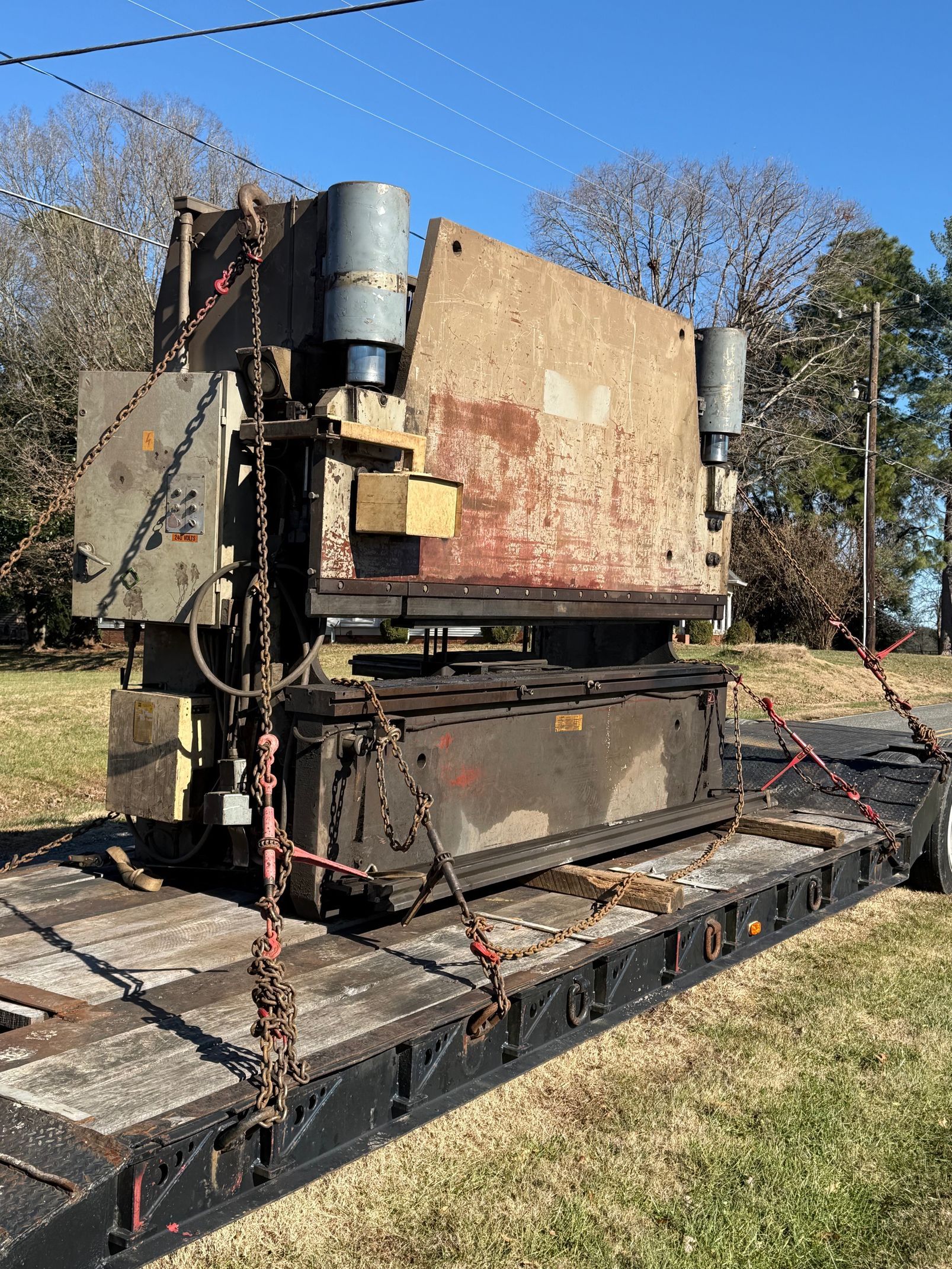 A large, industrial hydraulic press machine secured with chains on a flatbed trailer outdoors on a sunny day.