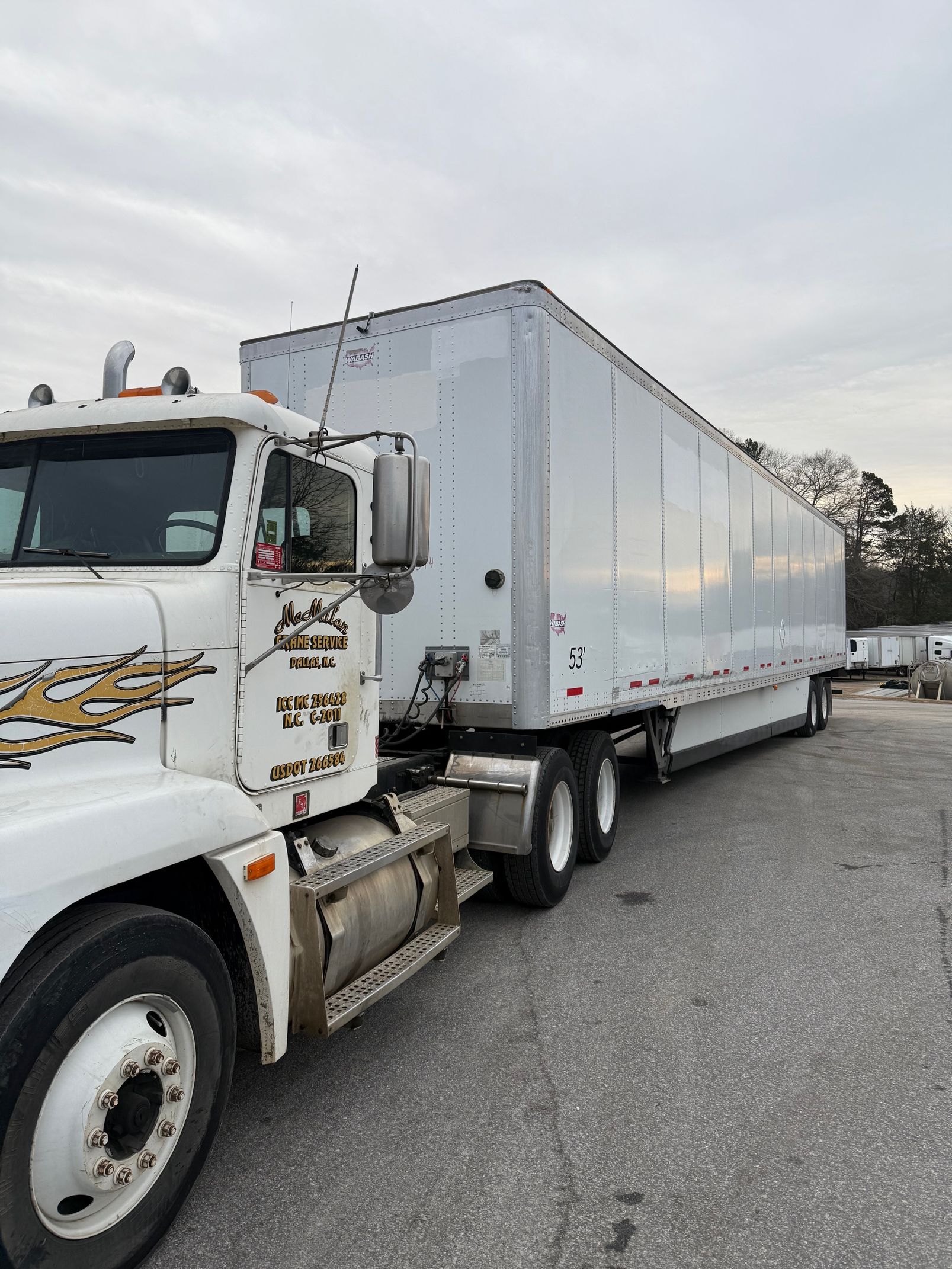 A white semi-truck with a long trailer parked on a gravel lot under a cloudy sky.