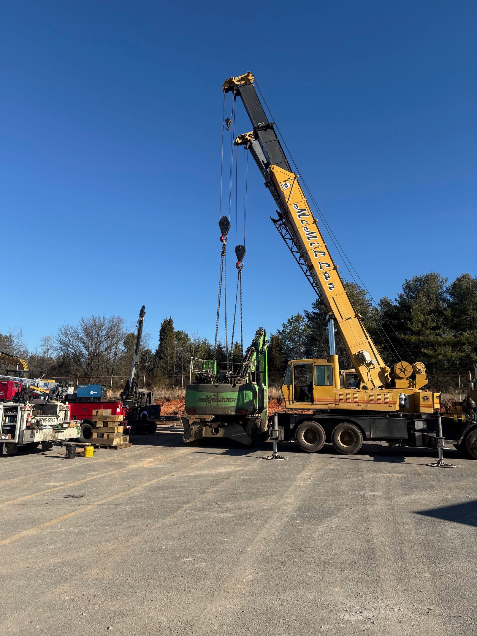 A yellow mobile crane stands in an outdoor lot, lifting equipment near other trucks and construction vehicles.