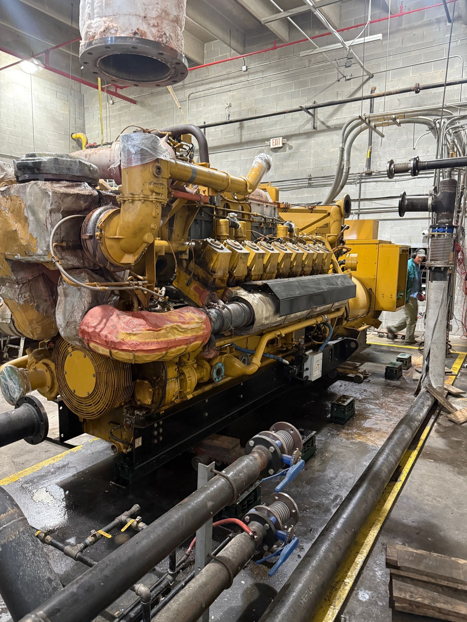 A large, yellow industrial engine sits in a workshop with connected pipes and exhaust components under a metal ceiling.