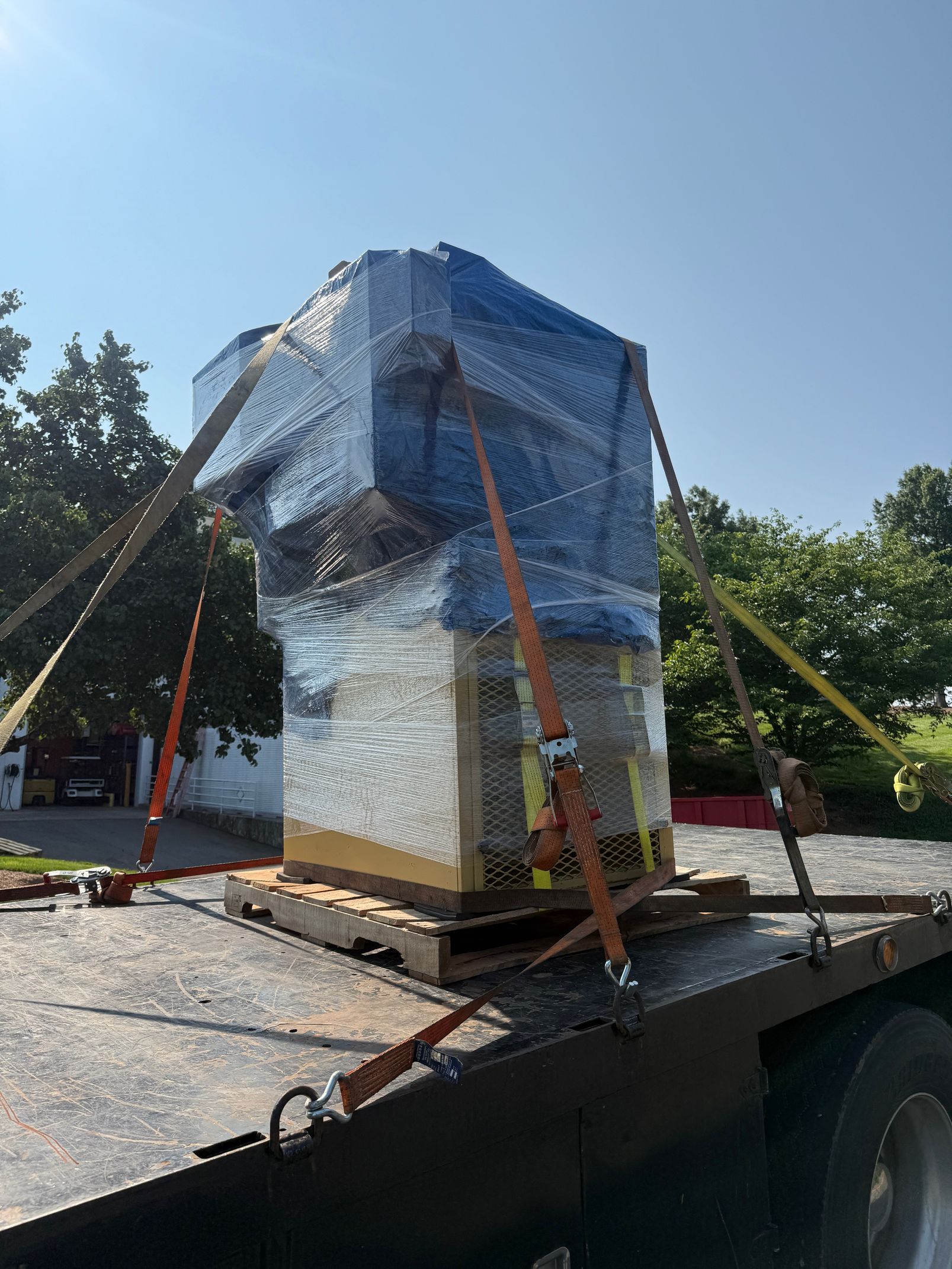 A large object wrapped in blue and white plastic, secured with orange straps on a flatbed trailer outdoors.