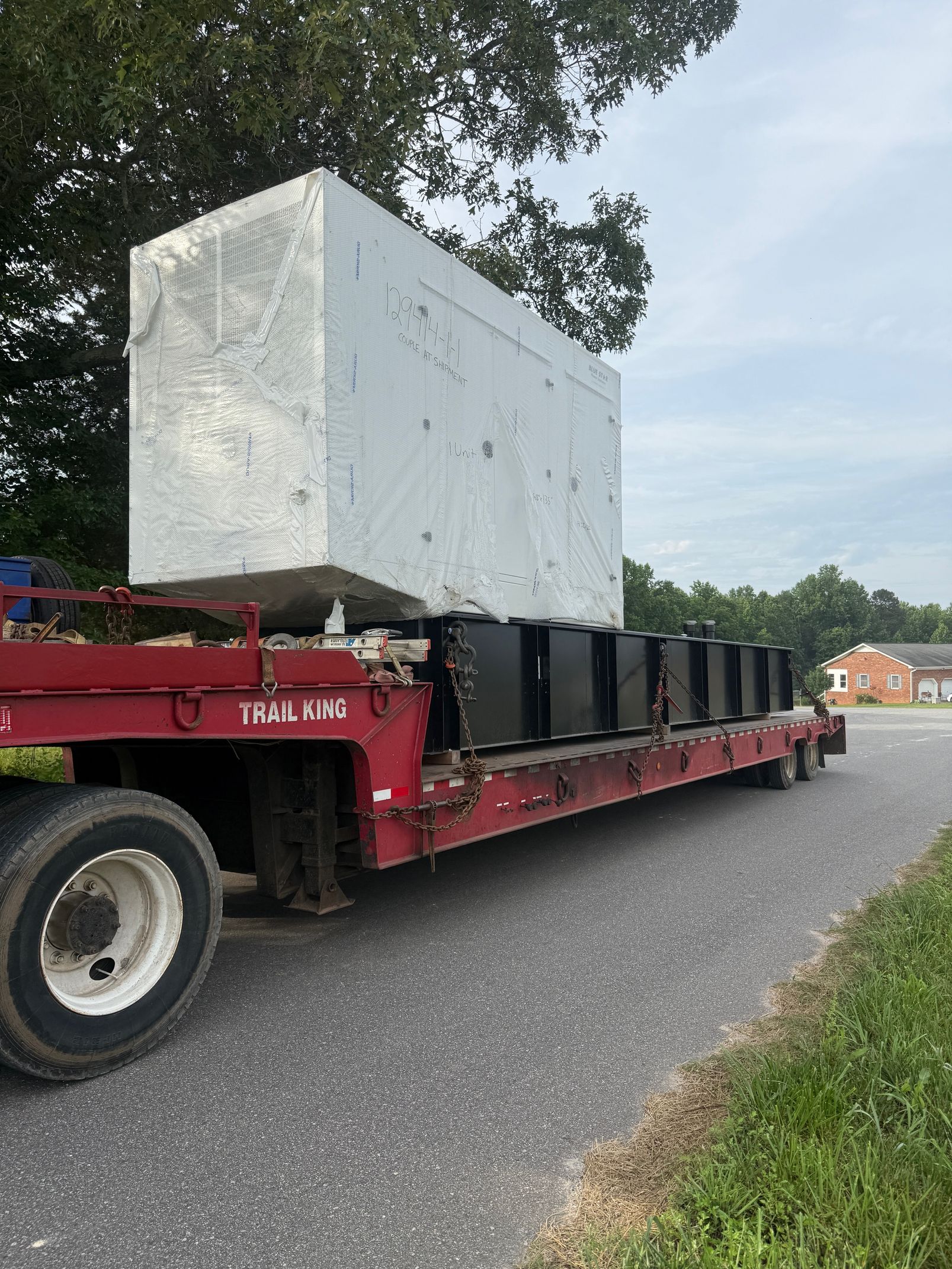 A large, rectangular concrete block sitting on a flatbed trailer towed by a red truck, parked on a roadside.
