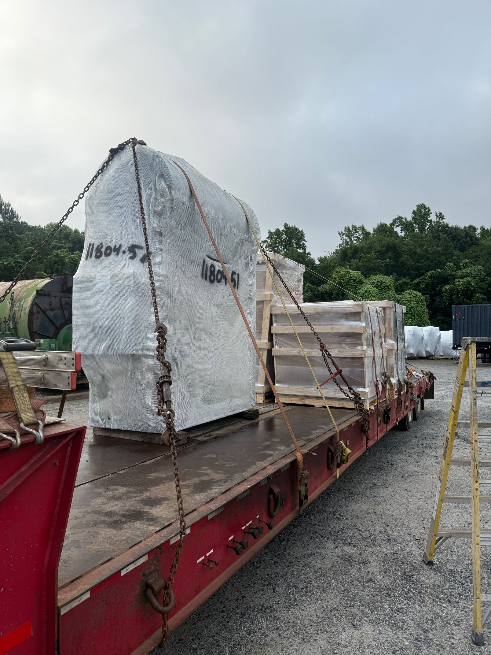 A flatbed trailer loaded with shrink-wrapped industrial equipment secured by chains at an outdoor storage lot.