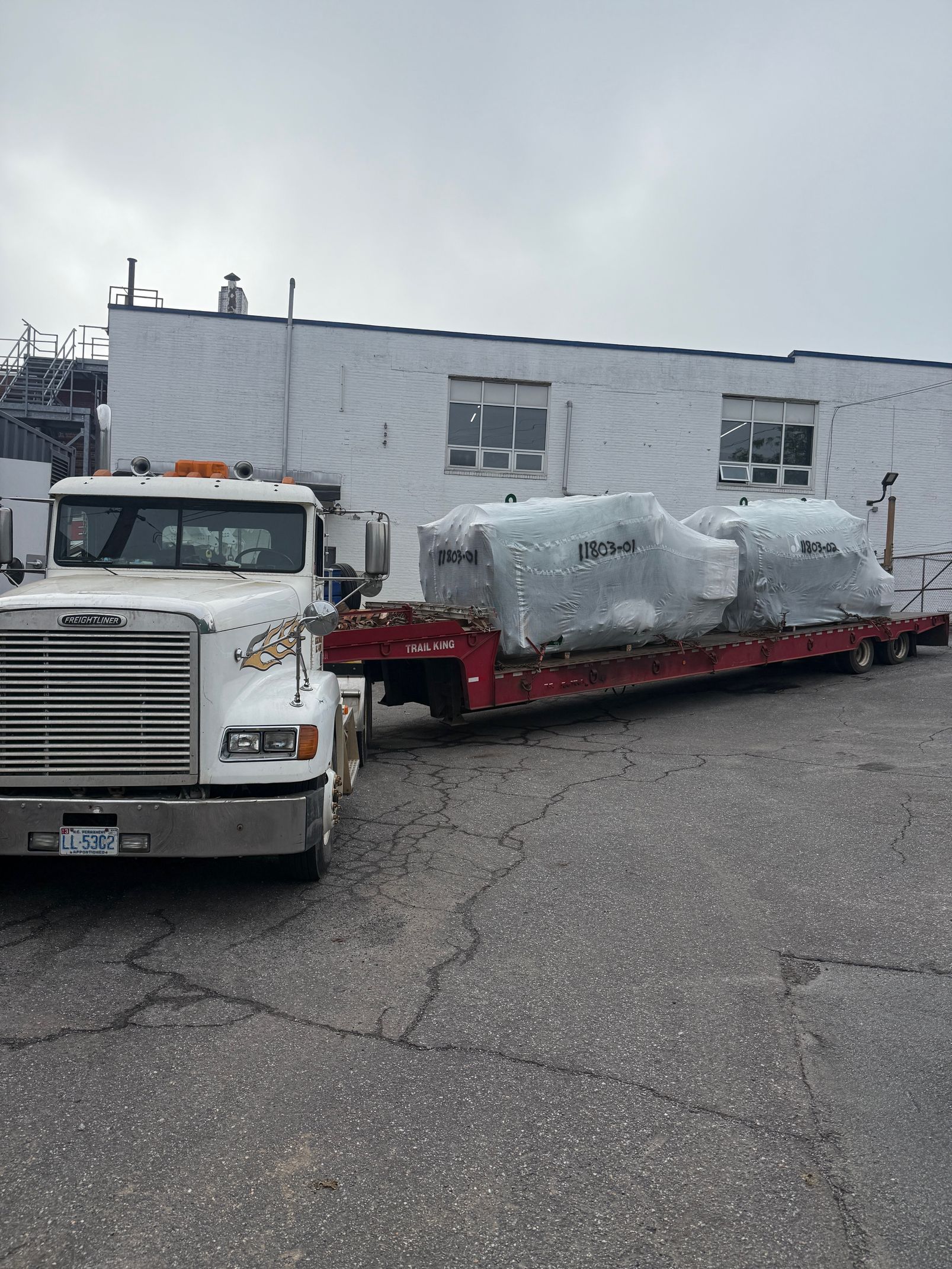 A white semi-truck parked in a gravel lot with two large, plastic-wrapped industrial machines loaded on a flatbed trailer.