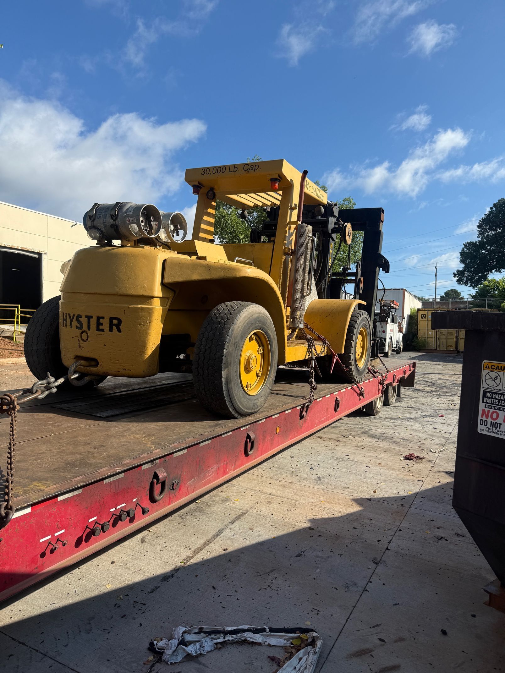 A large yellow Hyster forklift sits on a red flatbed trailer outdoors under a blue sky.