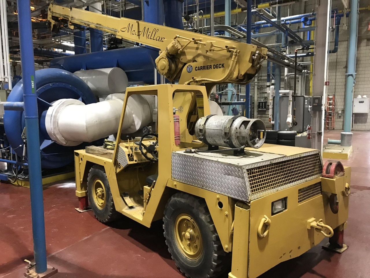 A yellow industrial crane truck parked indoors on a dark floor near large machinery and white pipes.