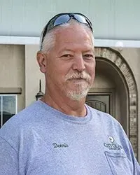 A man wearing sunglasses and a grey shirt is standing in front of a building.