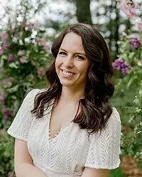 A woman in a white dress is smiling in front of purple flowers.
