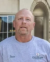 A man with a beard is wearing a grey shirt and standing in front of a building.