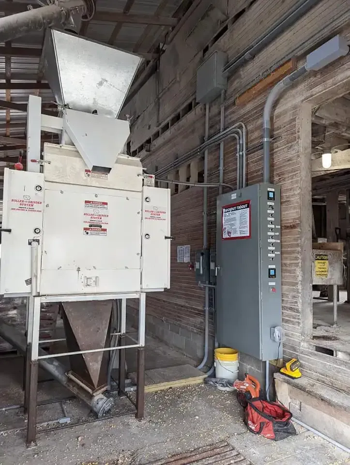 A white, industrial milling machine with a hopper stands next to a gray electrical panel in a brick-walled facility.