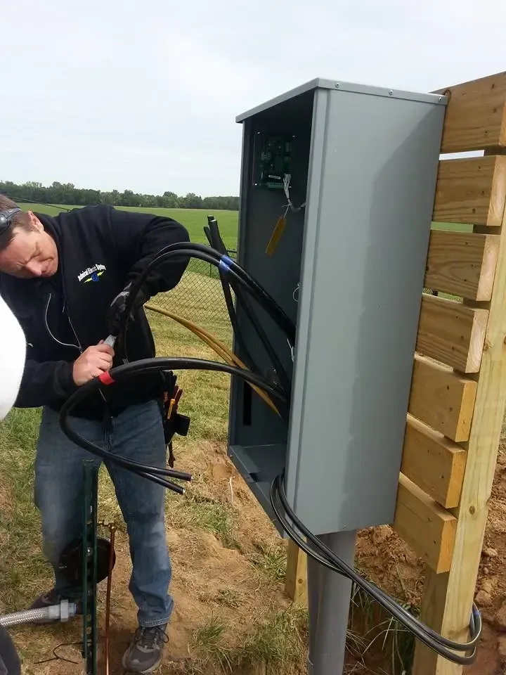 A technician installs electrical wiring into a grey outdoor utility box mounted on a wooden post in an open field.