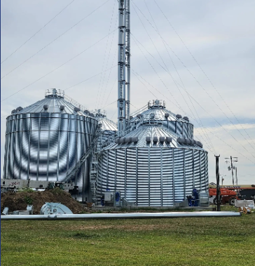 Two large corrugated metal grain bins with a tall grain elevator leg set against a cloudy sky.