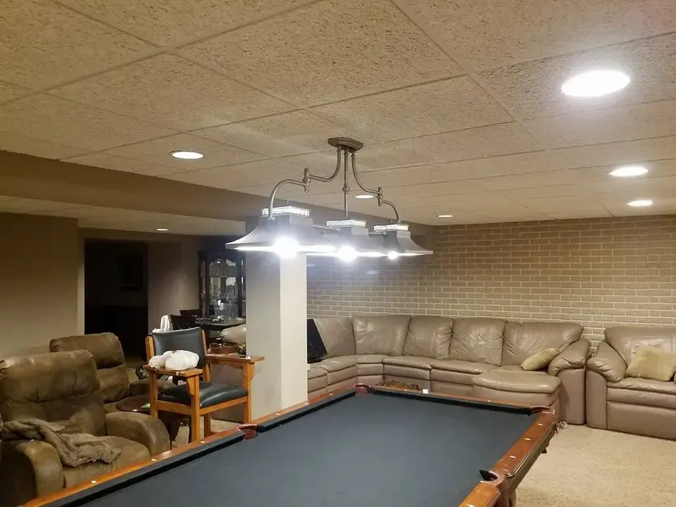 A finished basement featuring a pool table illuminated by a three-light chandelier, surrounded by tan leather seating.
