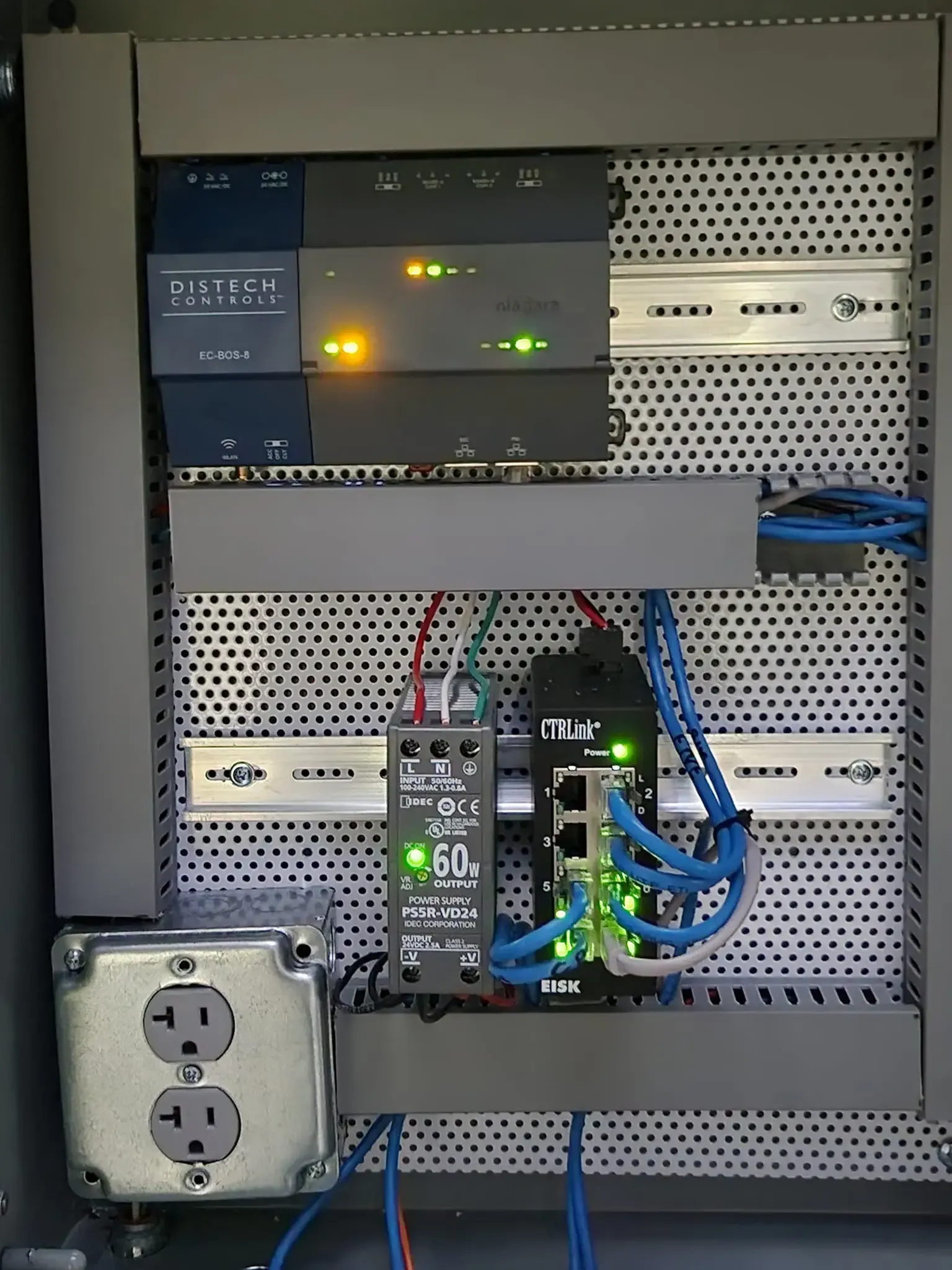 Industrial control cabinet containing a gray PLC unit, power supply, and network switch with status lights on a DIN rail.