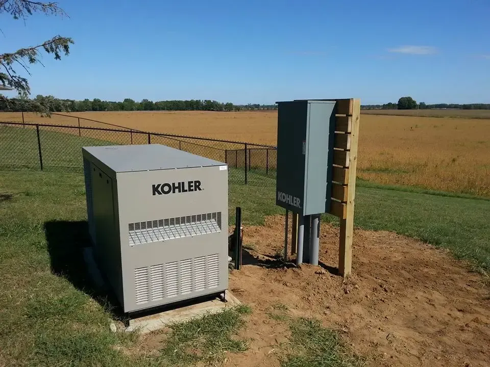 A grey Kohler backup generator and matching metal utility box stand on a dirt patch beside a fence in a grassy field.