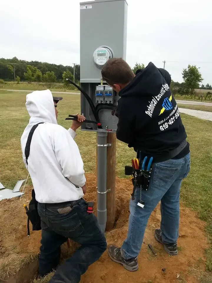 Two technicians install wiring in an outdoor electrical meter box mounted on a wooden post in an open field.