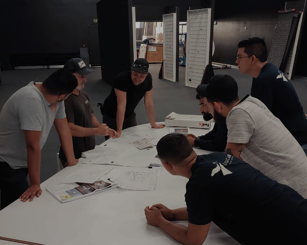 Group of men leaning over a white table, examining papers and objects in an indoor setting.