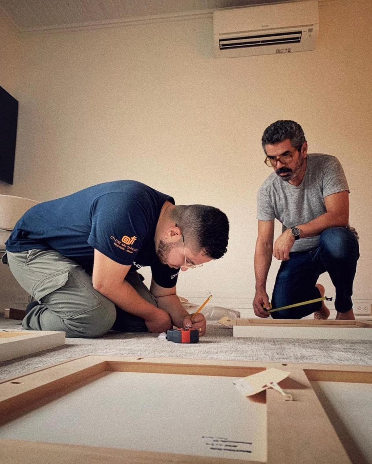 Two people working on a wooden frame on a floor. One is kneeling and marking, the other measuring. Indoors.