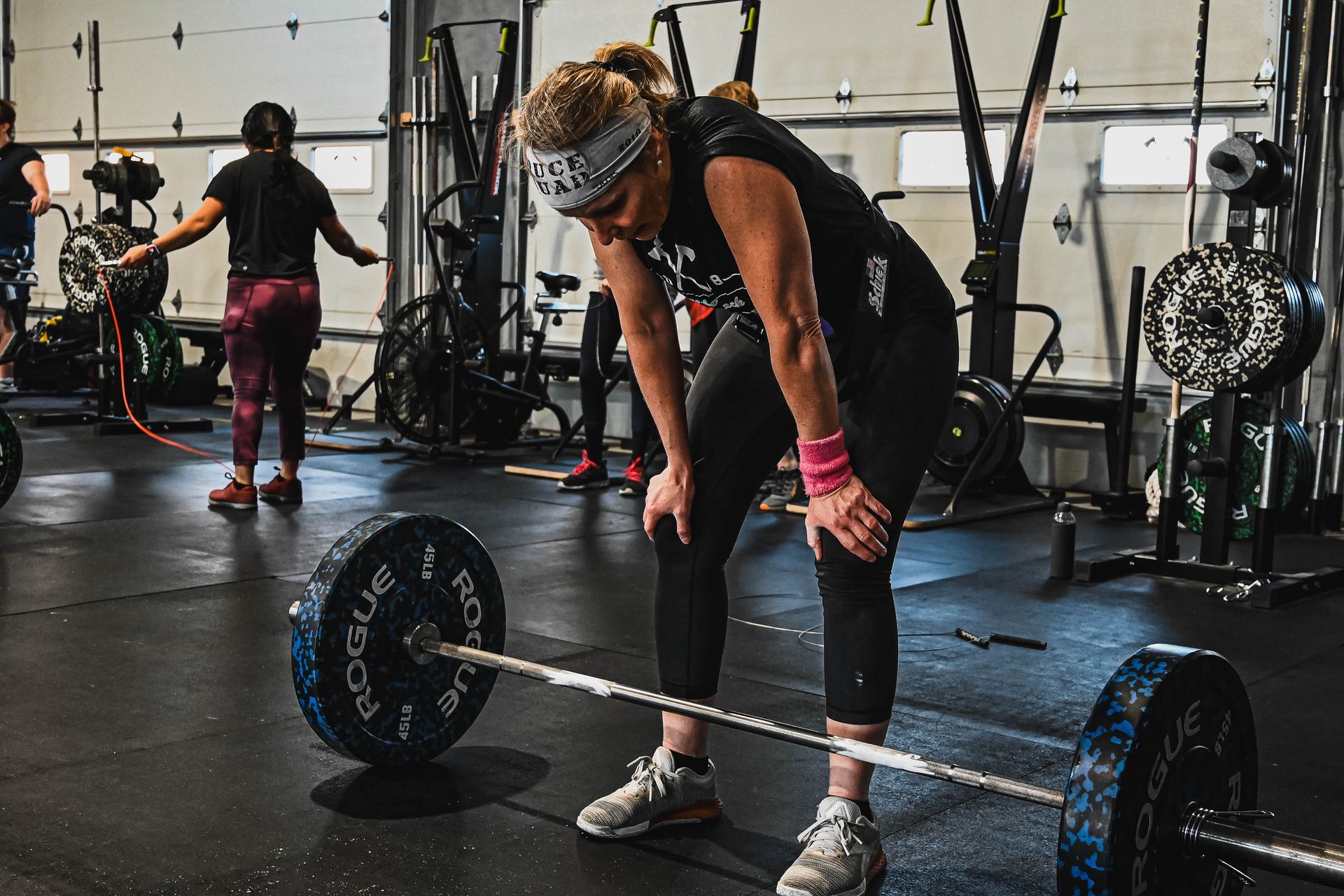 Woman resting hands on knees after lifting a barbell in a gym. Other people are working out in the background.