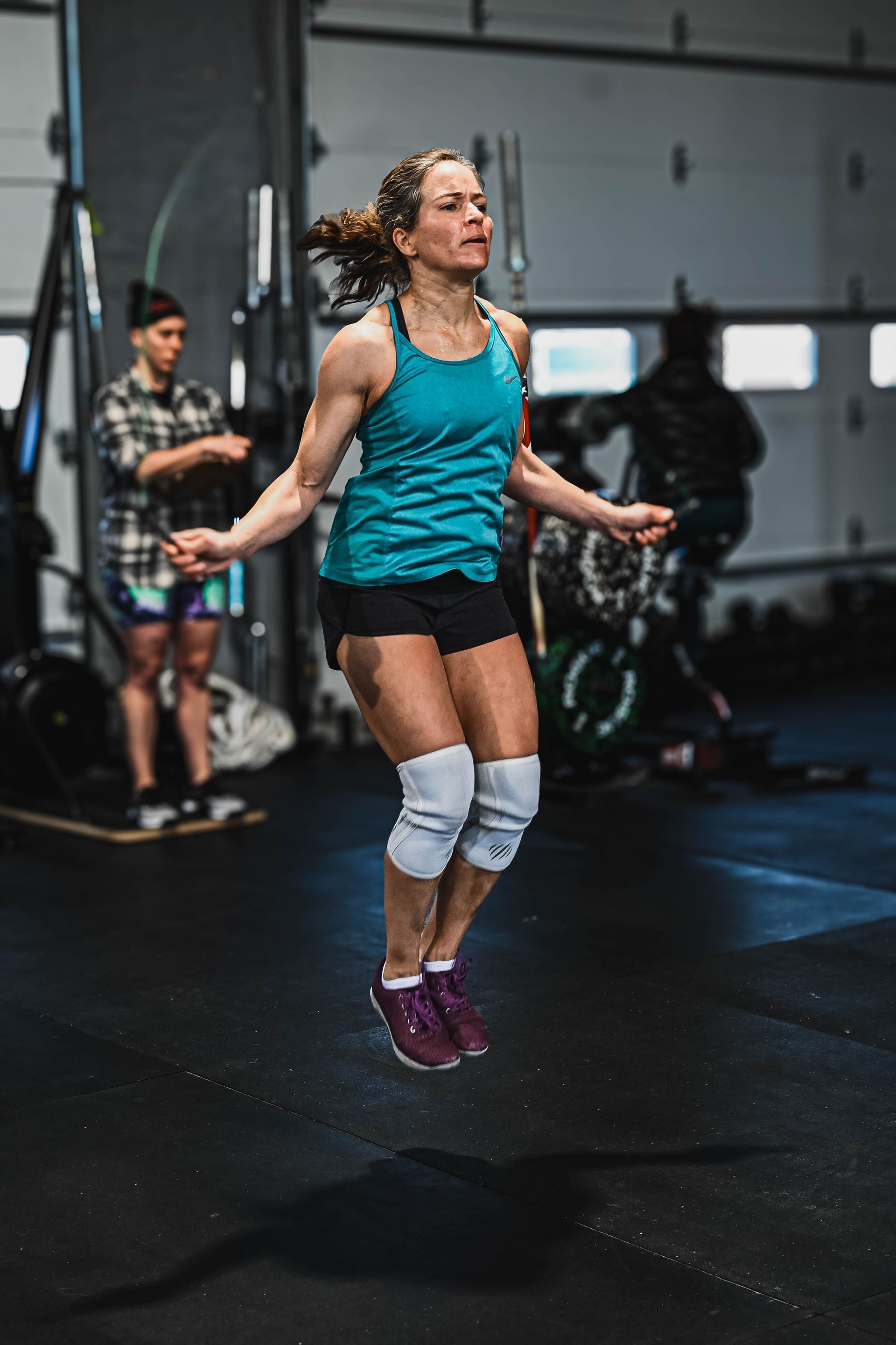 Woman jumps rope in gym, wearing teal tank, black shorts, knee pads; another person watches.