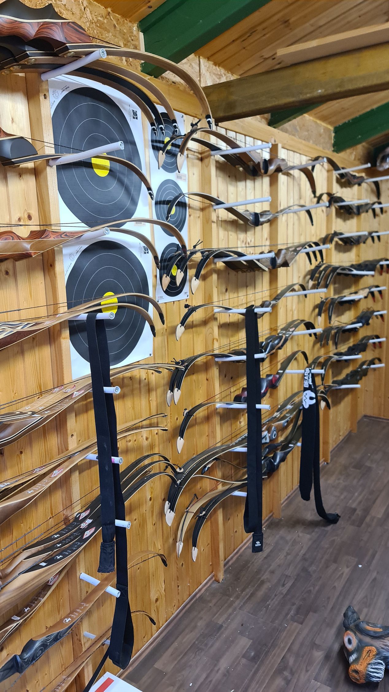 In einem holzgetäfelten Gebäude sind Bögen auf Holzregalen ausgestellt, im Hintergrund sind Bogenschießscheiben zu sehen.