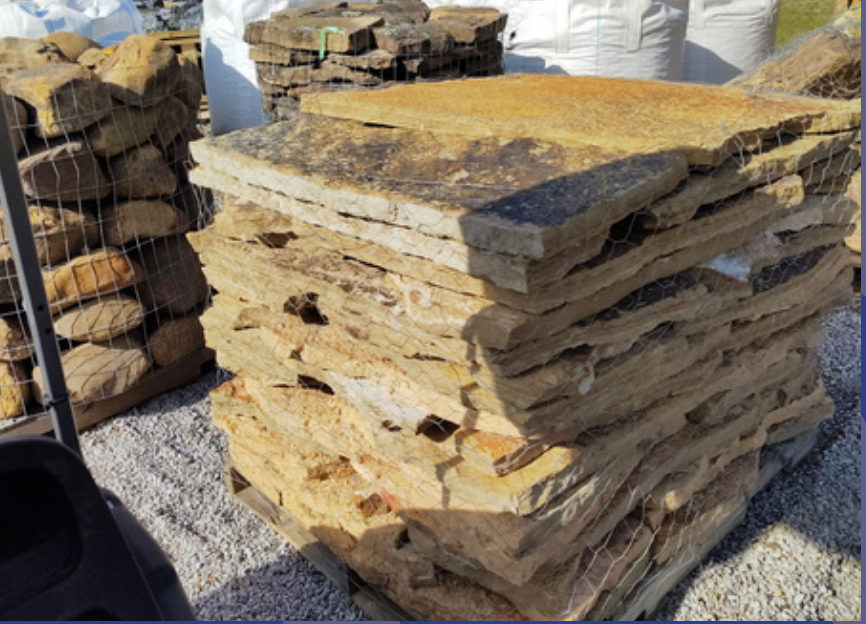 A stacked pallet of flat, rectangular tan and brown natural stone pavers in an outdoor yard setting.