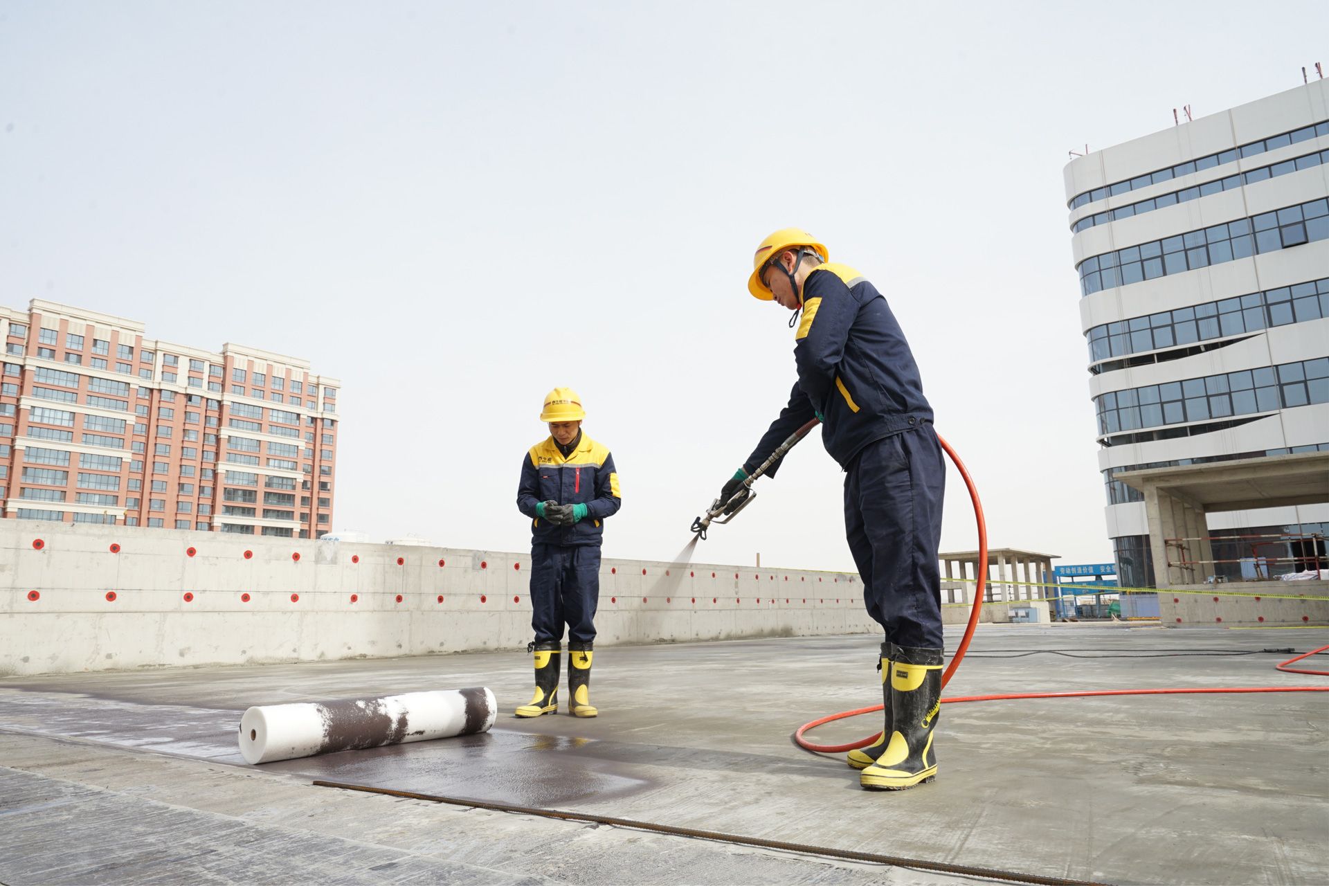Two construction workers in safety gear using a power hose on a rooftop worksite.
