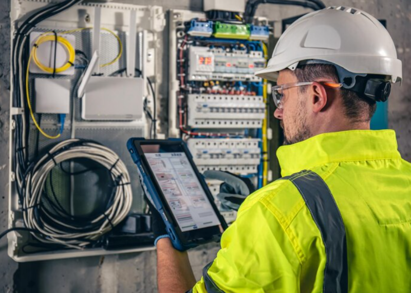 A technician in a high-visibility jacket and hard hat uses a tablet to inspect wiring inside an open electrical cabinet.