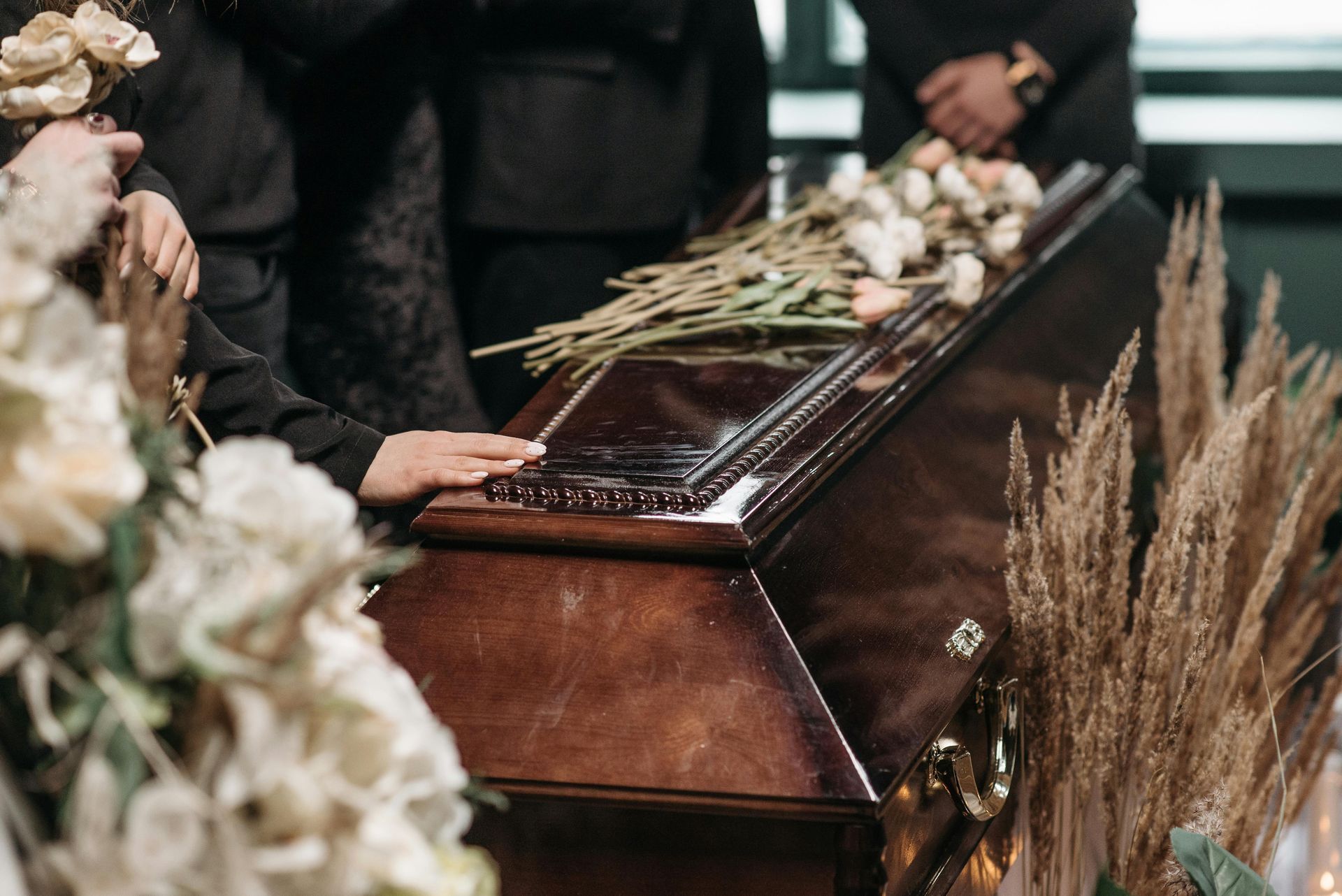 A hand rests on a closed, brown wooden casket adorned with flowers.