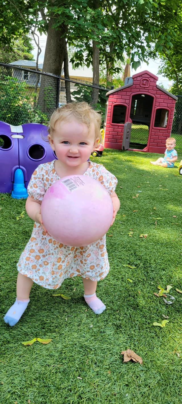 A little girl is standing in the grass holding a pink ball.
