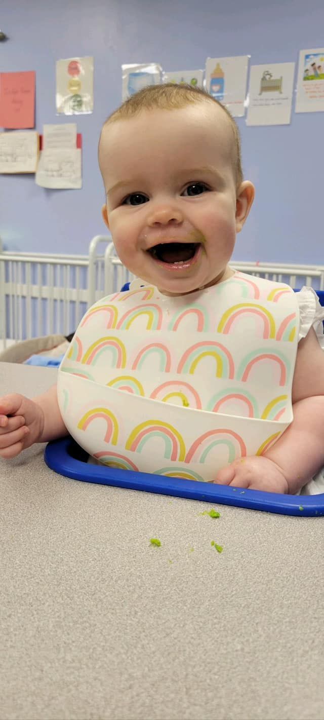 A baby wearing a bib is sitting in a high chair and smiling.