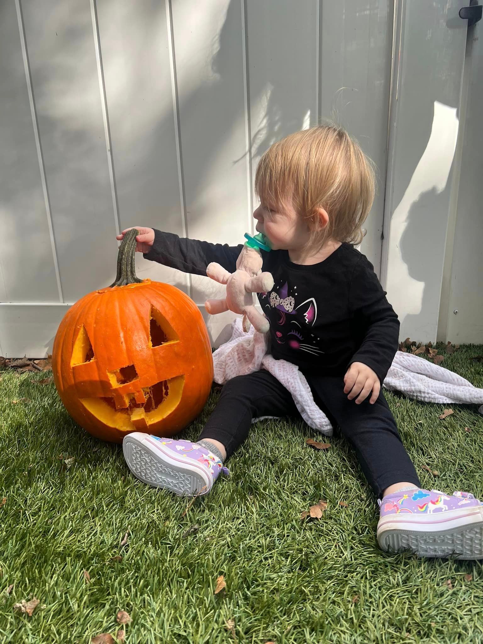A little girl is sitting on the grass next to a carved pumpkin.
