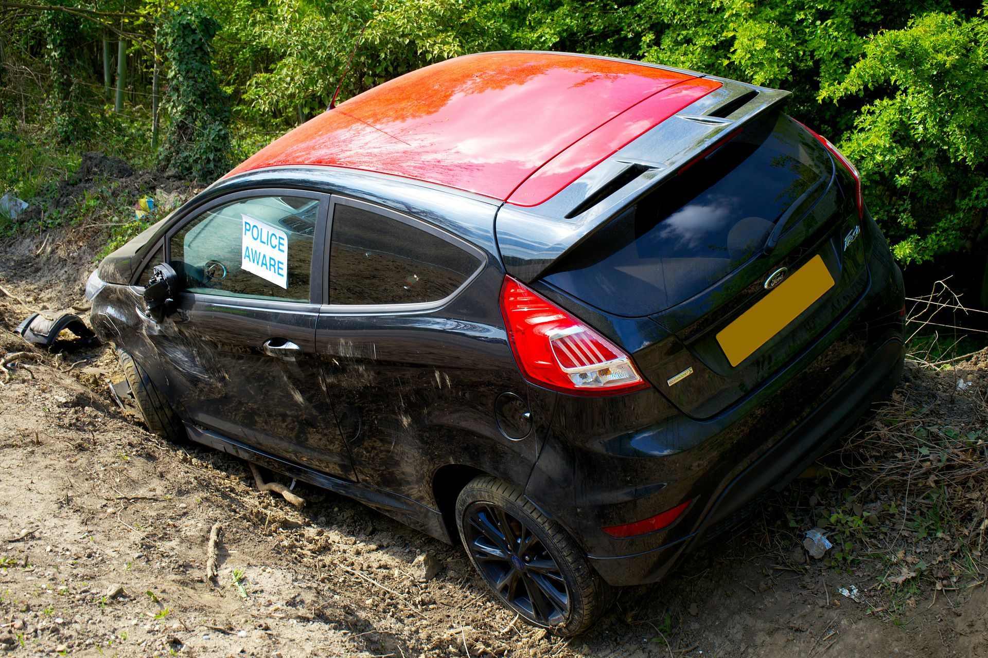 A black and red Ford Fiesta crashed into a muddy embankment surrounded by trees.