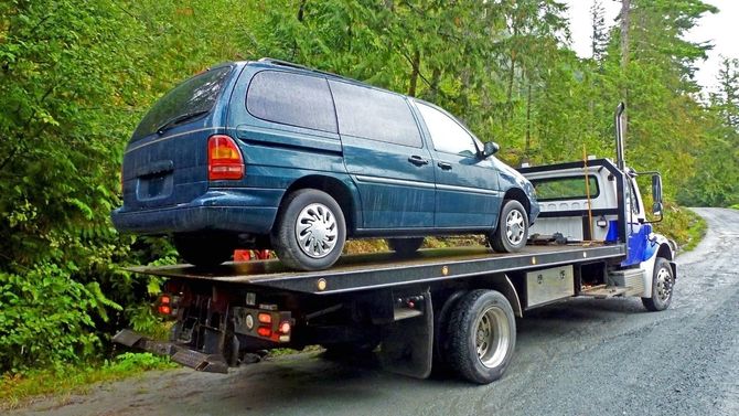 A tow truck uses a crane to lift a blue convertible, while a white sedan is parked on the street in front of them.