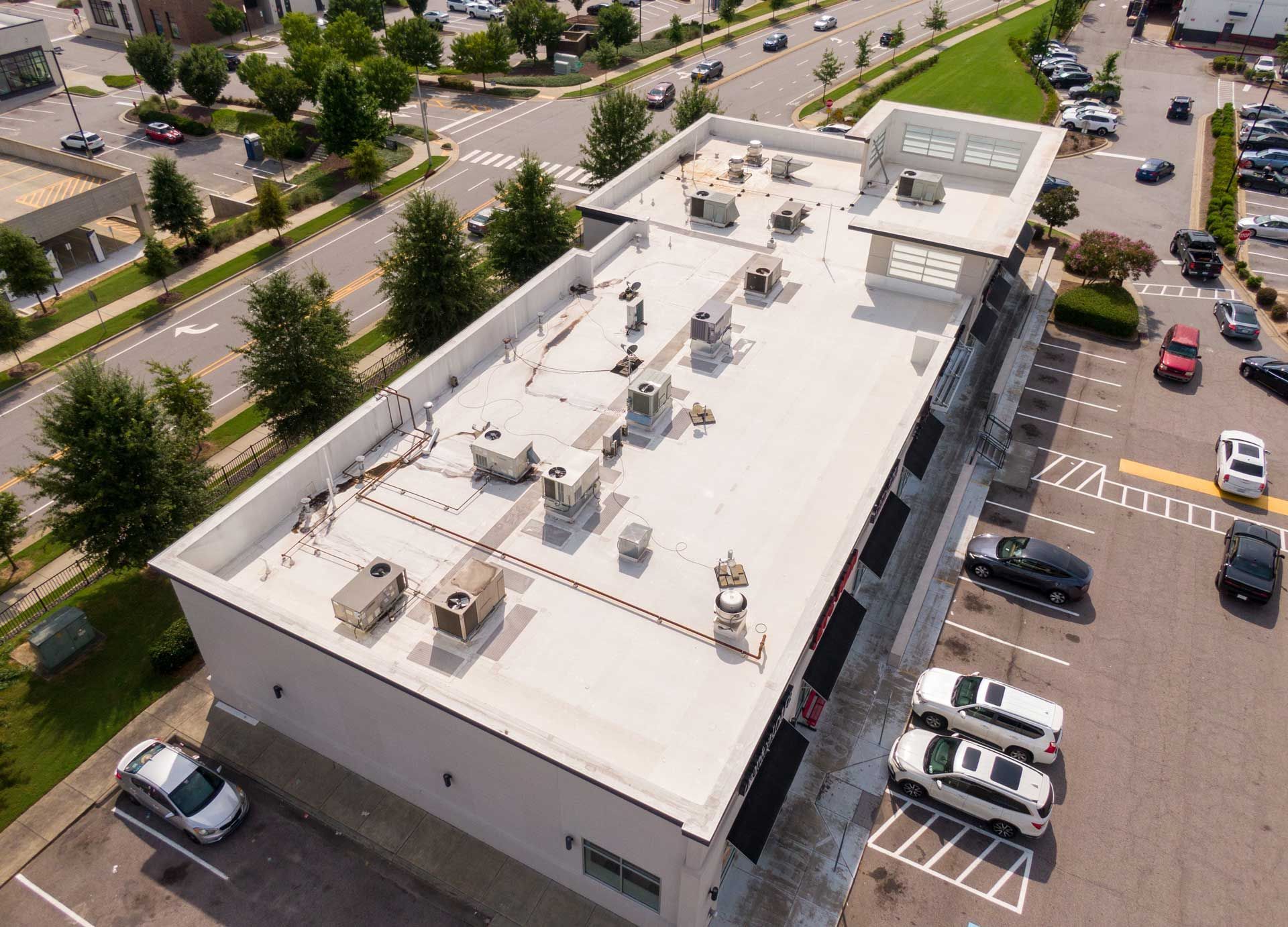 Overhead view of a building with a flat white roof, AC units, and parking lot with cars. Overhead view of a building with a flat white roof, AC units, and parking lot with cars.