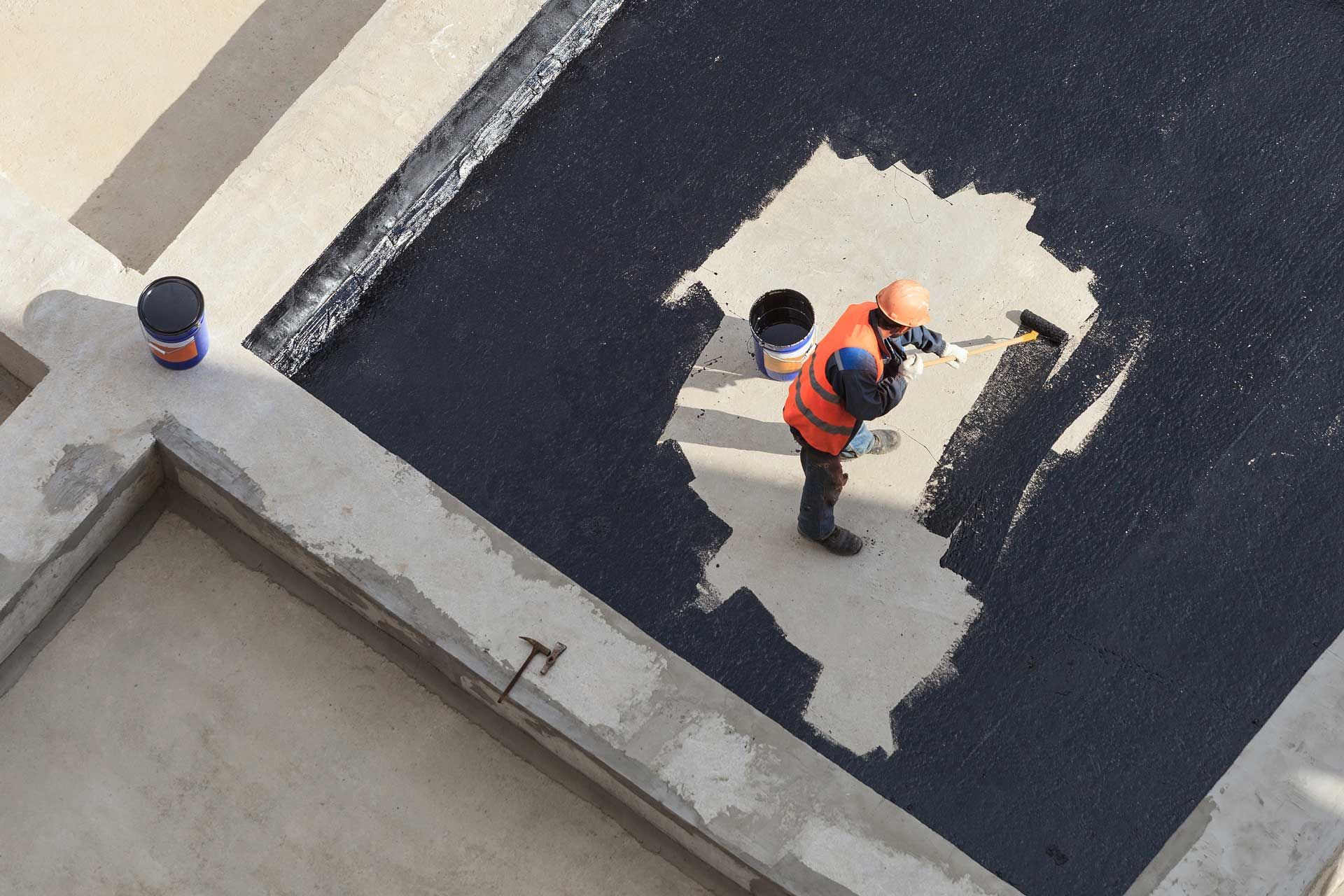 Construction worker applying sealant on a concrete surface, wearing an orange vest and hardhat.