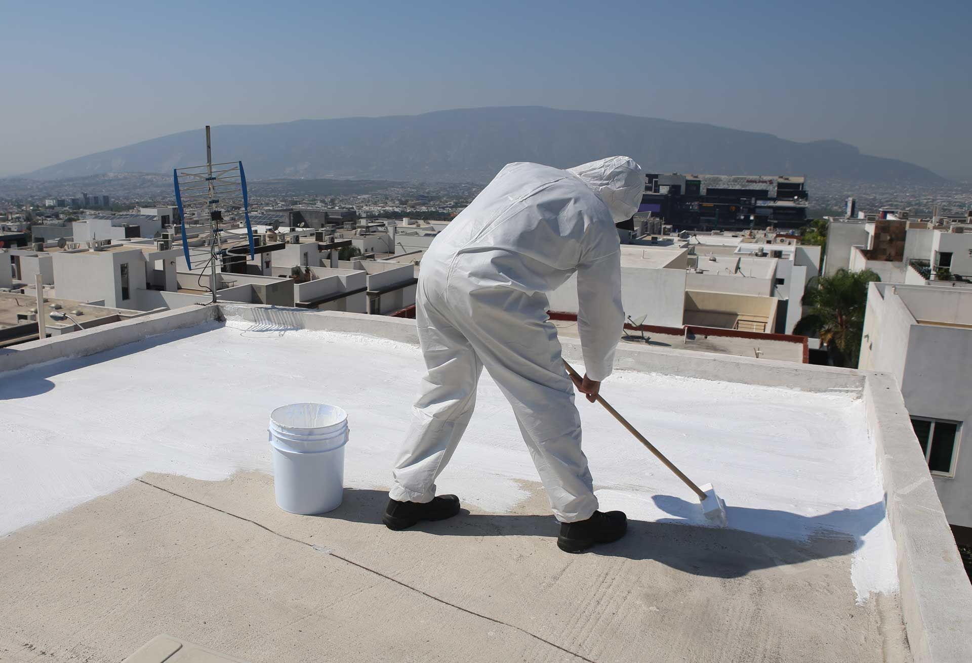 Person in protective suit applying white coating to a flat rooftop with a roller. City and mountain background.