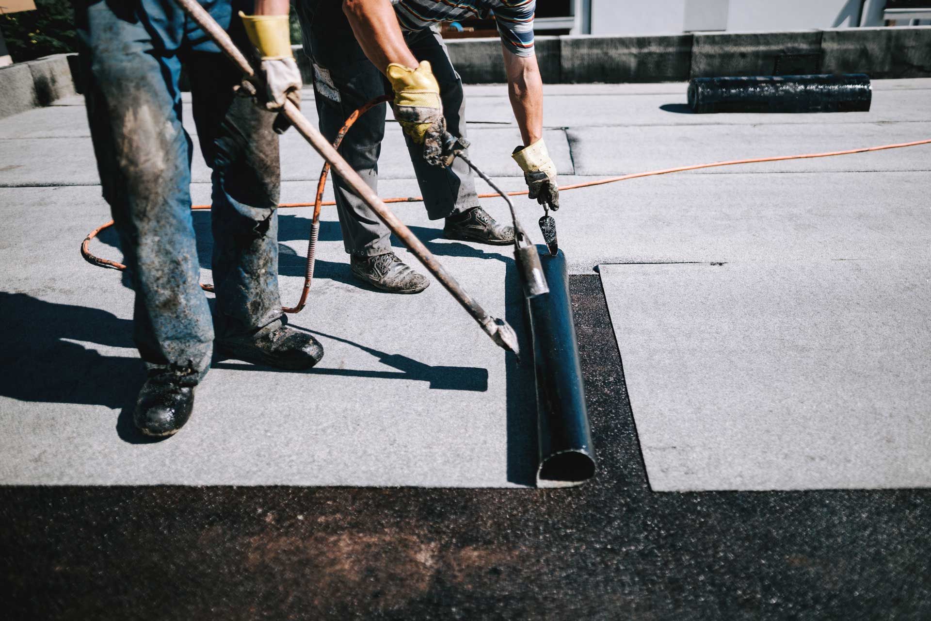 Two workers using a torch to apply black roofing material on a flat roof, wearing gloves and boots. Two workers using a torch to apply black roofing material on a flat roof, wearing gloves and boots.