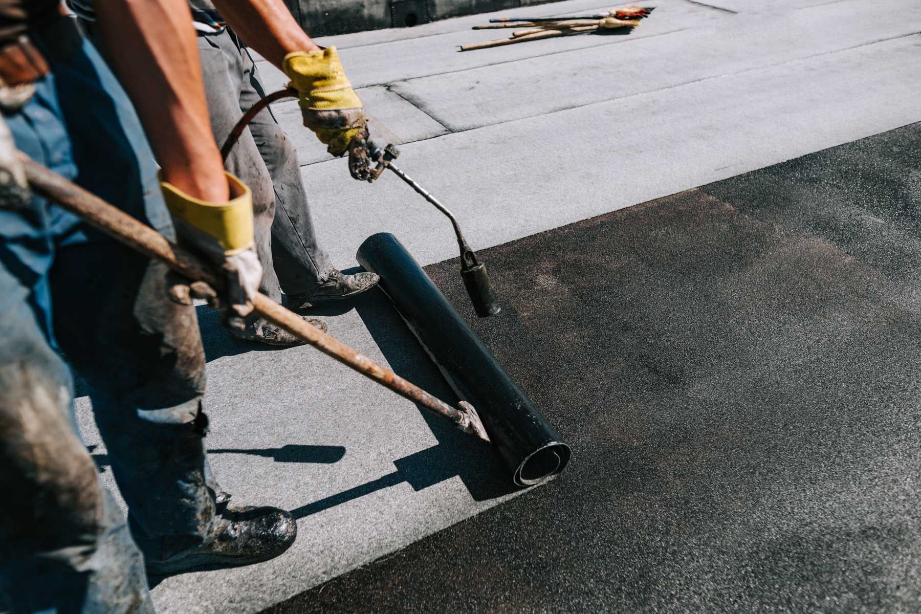 Workers installing roofing material on a flat surface using a torch and roller.