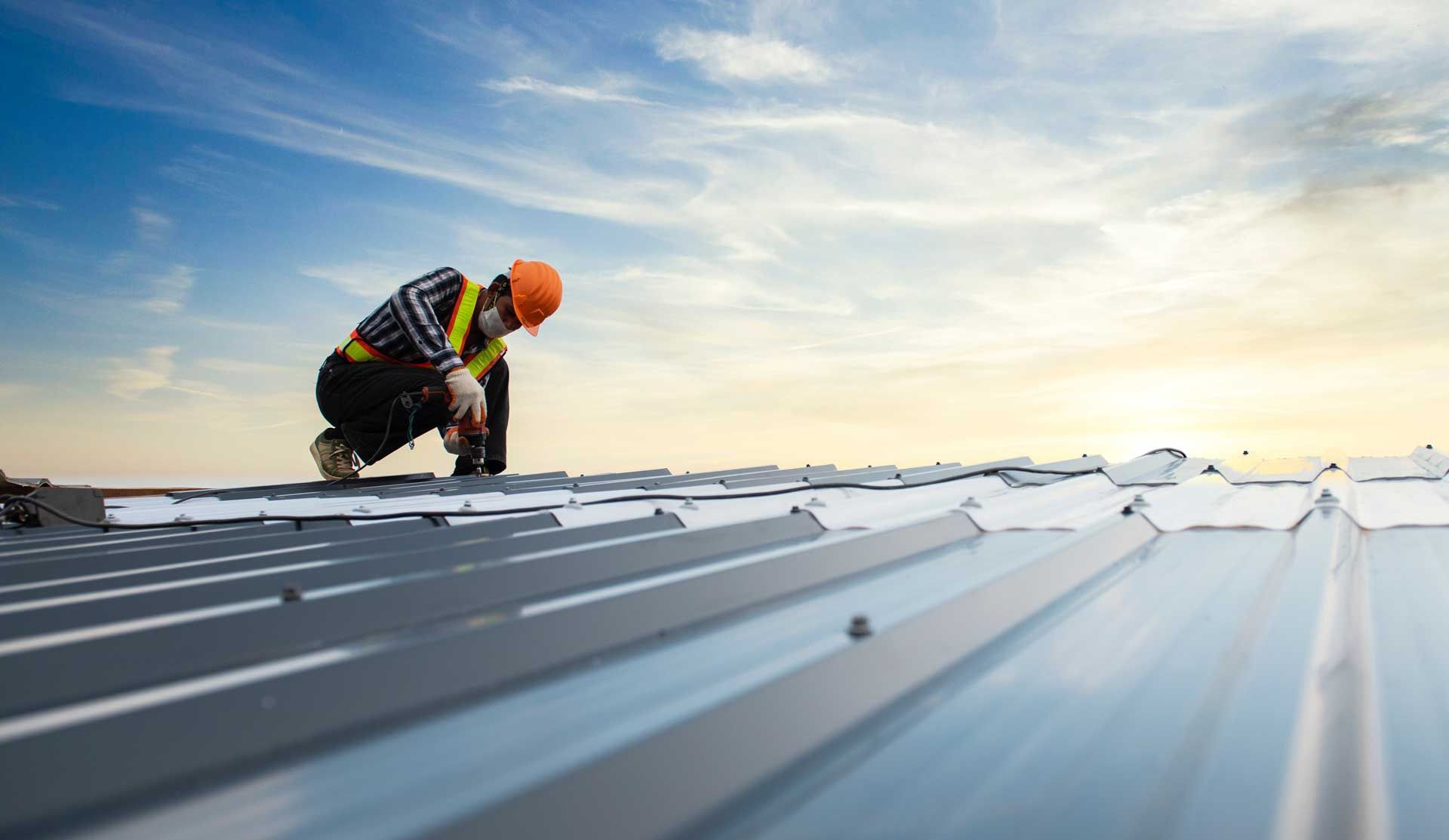Construction worker on a metal roof, wearing safety gear, inspecting the surface under a blue sky.