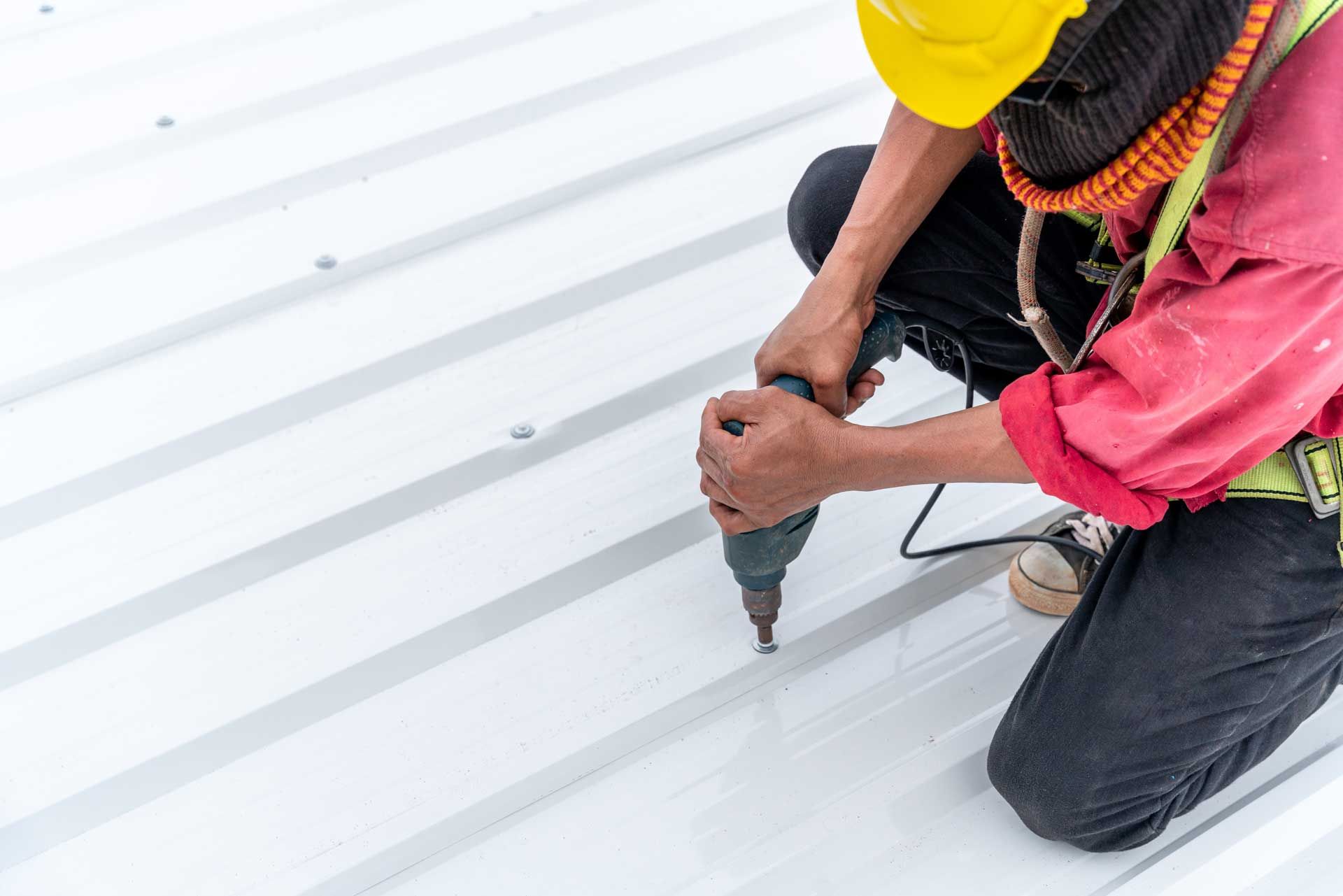 Person kneeling on a white metal roof, using a drill; wearing a hard hat and safety harness.