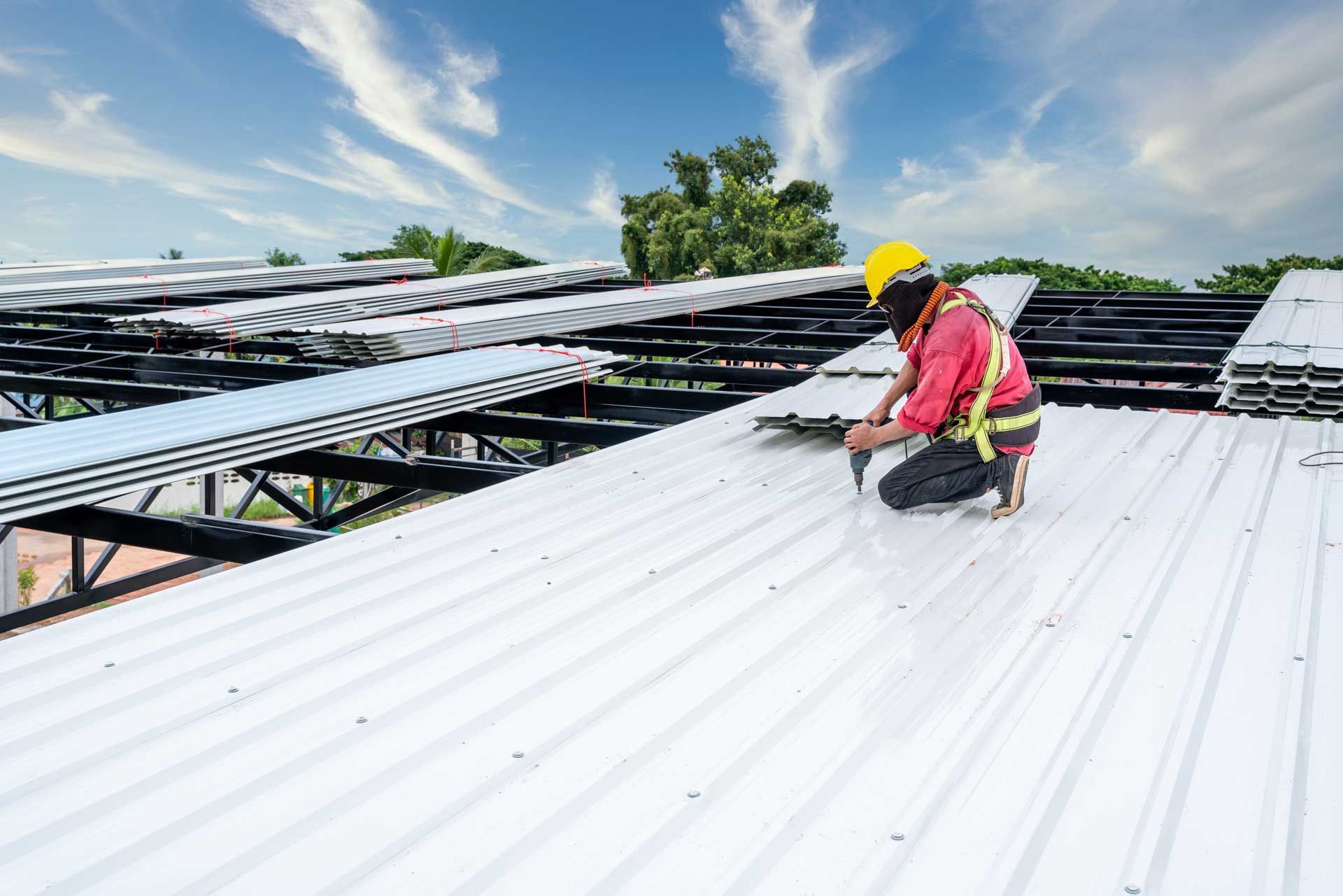 Roofer in safety gear installing metal roofing sheets on a building frame under a blue sky.