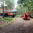 Orange utility truck and wood chipper in a yard near a cabin in a wooded area.