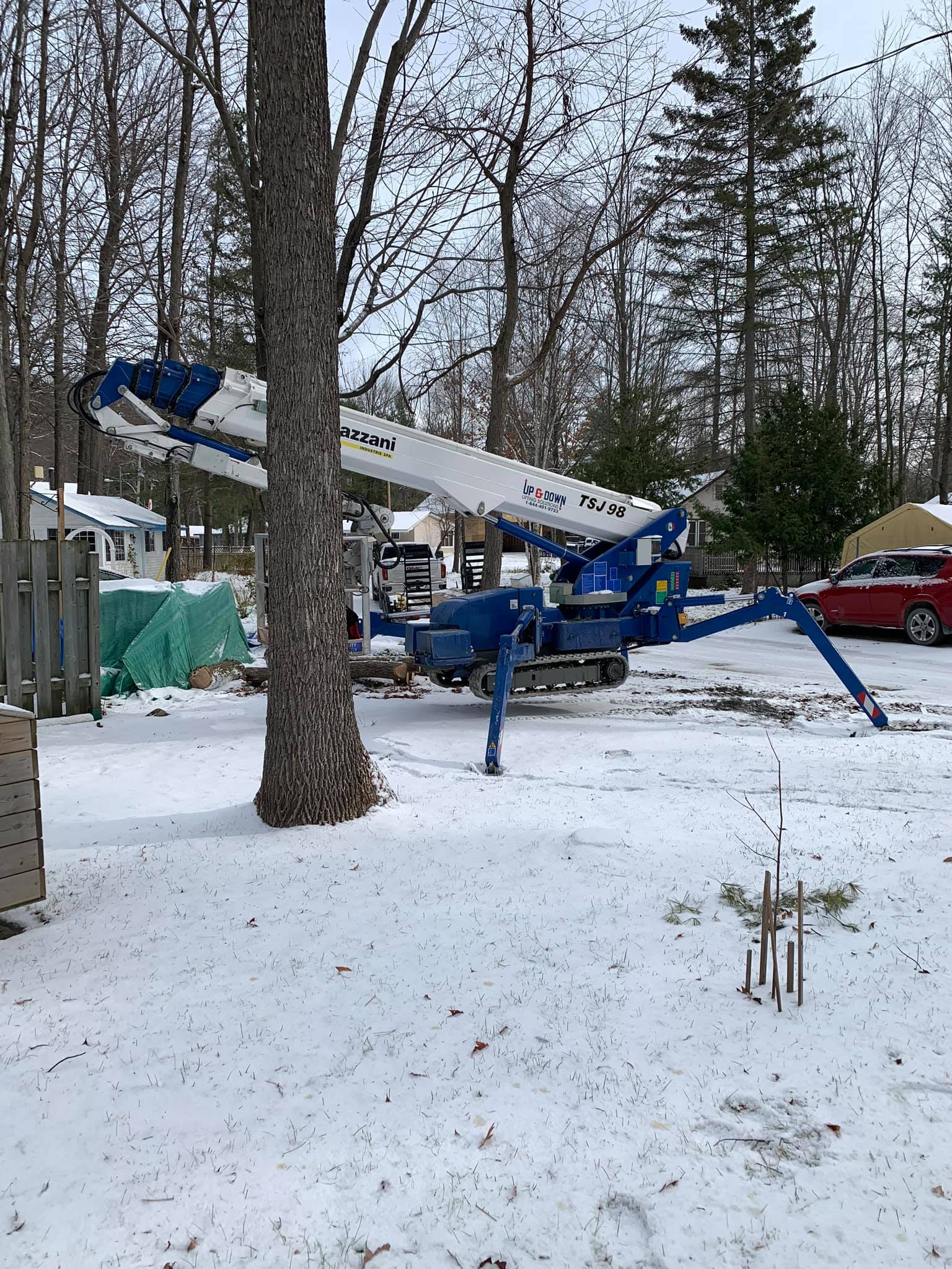 Blue lift machine with legs extended near a tree in a snowy yard.