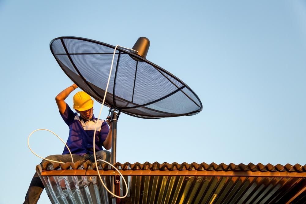 A Man Is Sitting on Top of A Roof Fixing a Satellite Dish — Peter Gow Electrical in Lismore, NSW